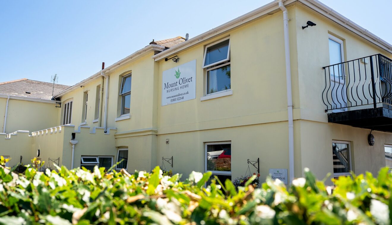 Mount Olivet Nursing Home's front elevation, with the reception sign and a flowering hedge in the foreground